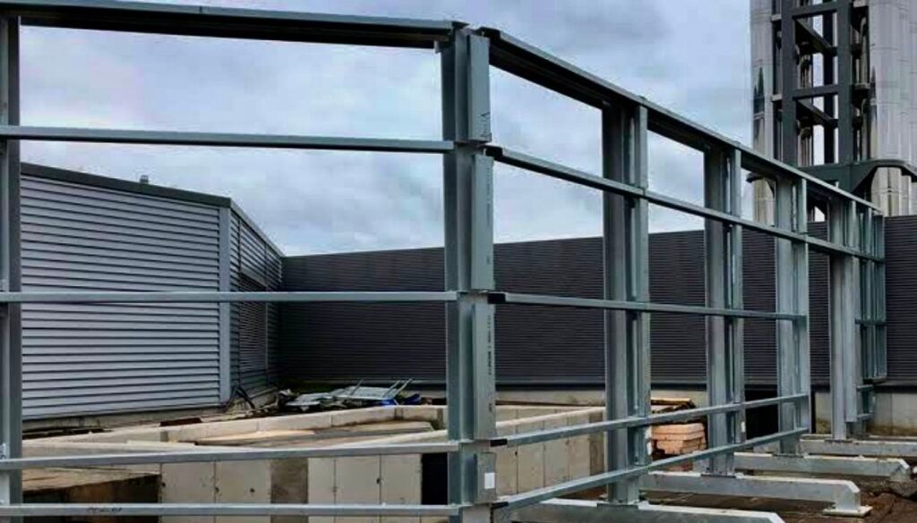 Image Showing A Construction Site With A Steel Framework In Progress. The Area Is Surrounded By Corrugated Metal Walls And Concrete Structures.
